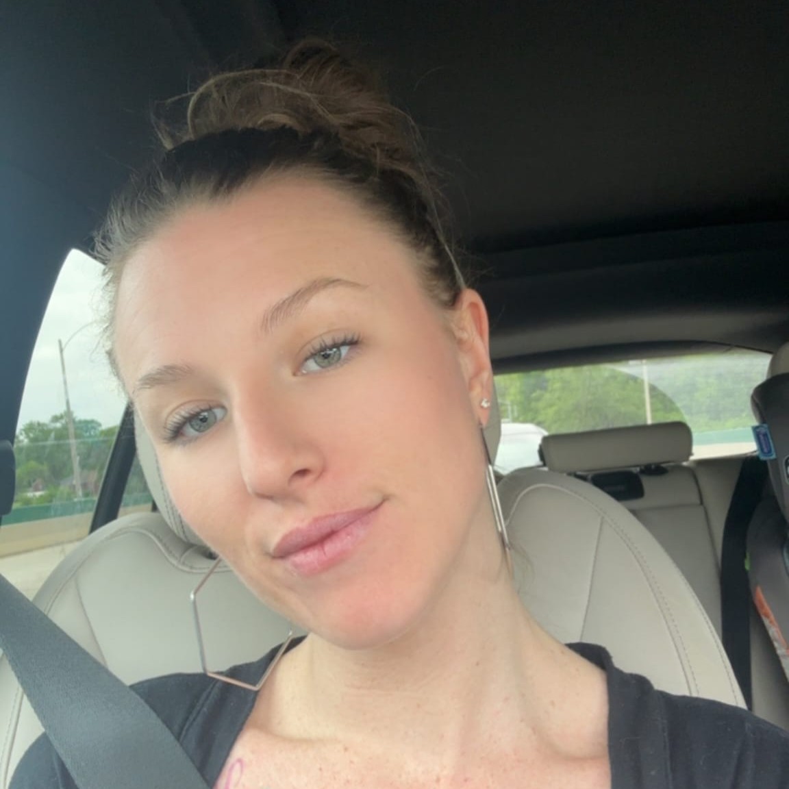 Close-up portrait of a person with hair in a bun seated in a car wearing a seat belt.