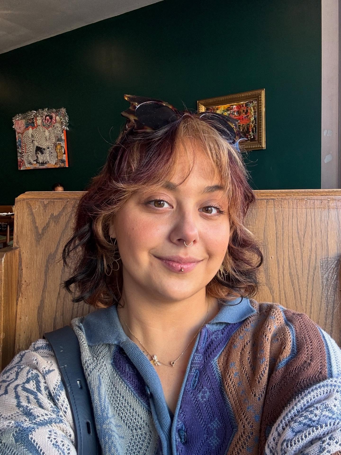 Smiling person with sunglasses on their head, septum ring, and colorful knit clothing seated before framed art.