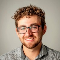 Smiling curly-haired man wearing rectangular glasses and a checked shirt against a plain gray background.