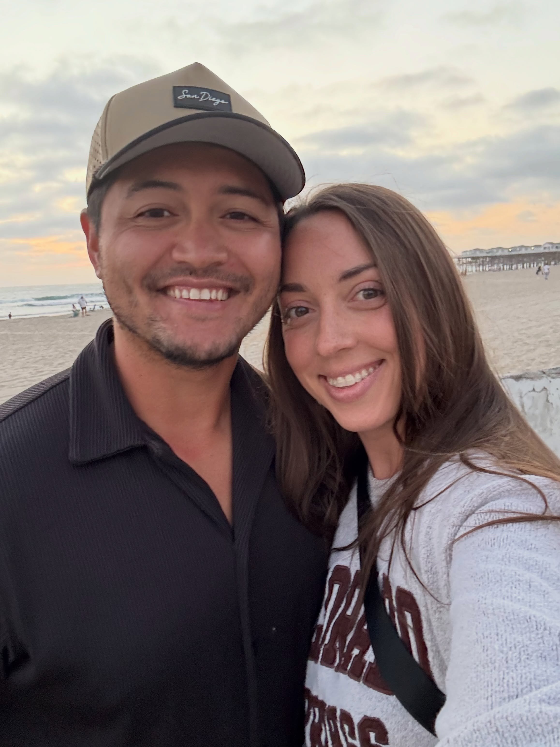 Two smiling adults take a close-up selfie on a beach at sunset, with a pier and ocean behind them.